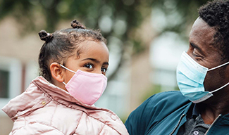 A man holds a young girl in his arms, and both are wearing COVID-19 face coverings.